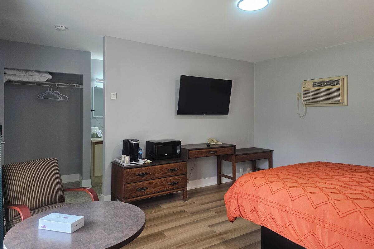 A hotel room with a bed, a desk with a chair, a television mounted on the wall, a wardrobe, and a small table with a box on it.