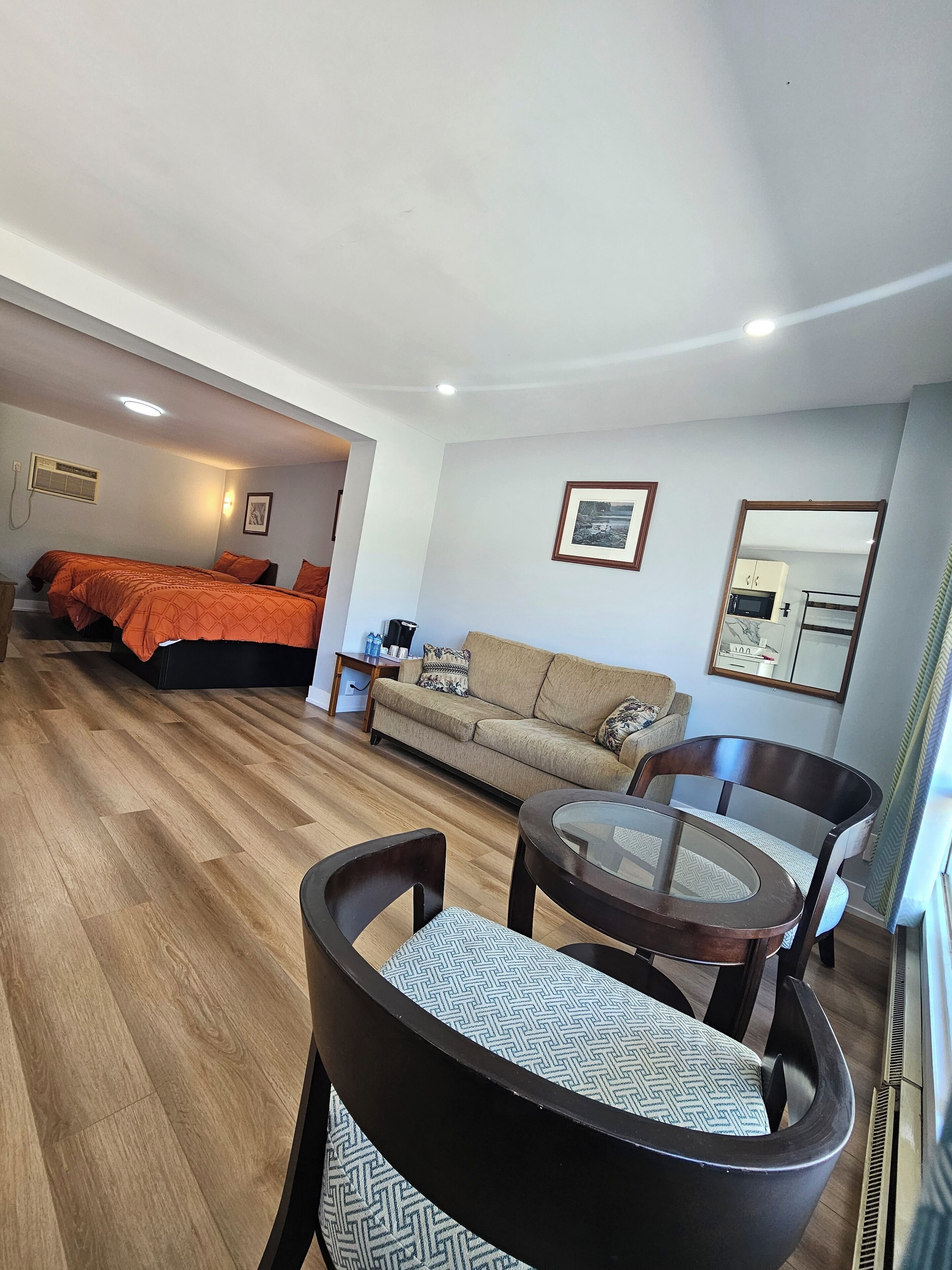 A hotel room with a bed, a sofa, a dining table with chairs, a kitchen area, and a mirror.