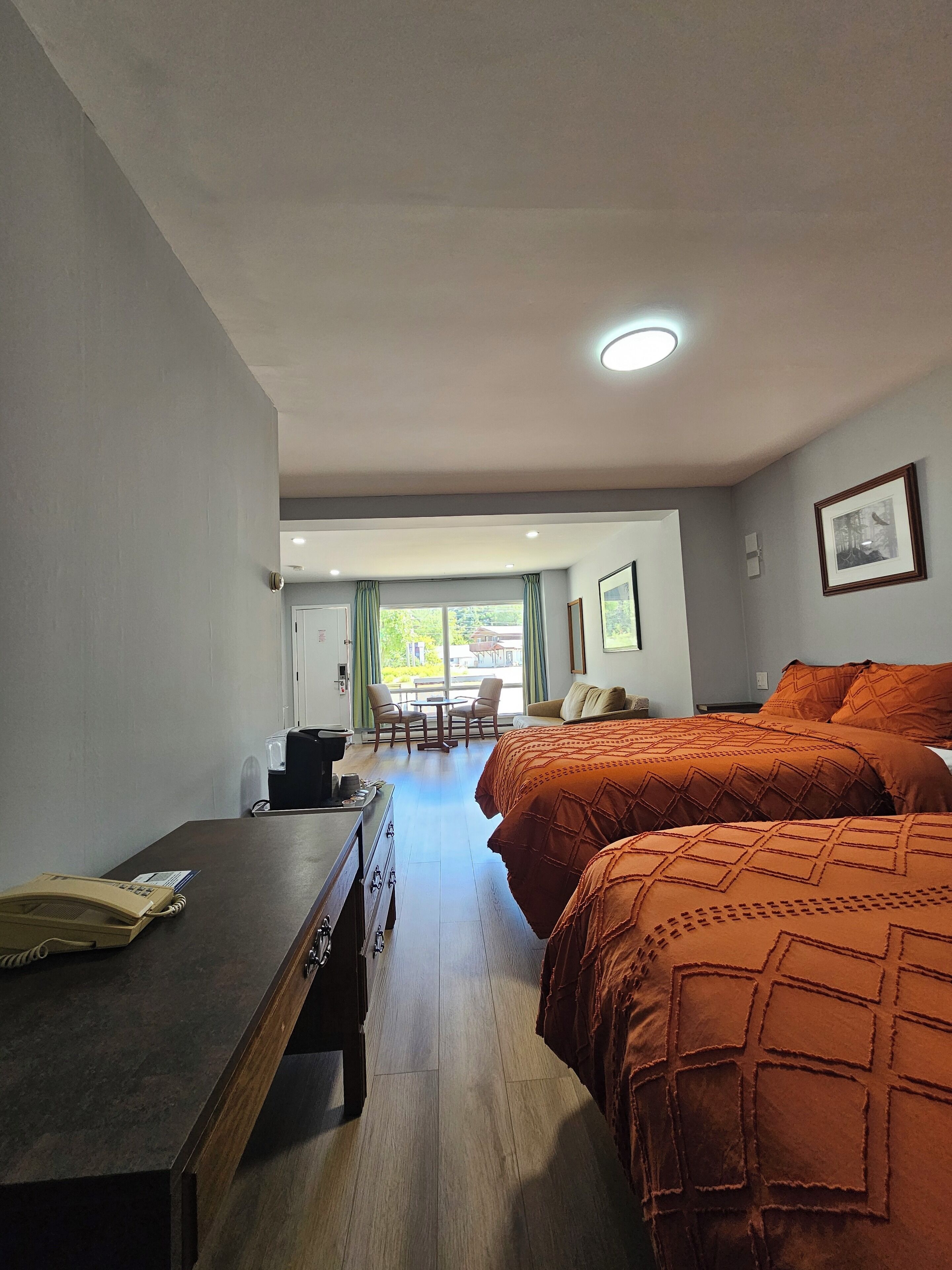 Une chambre d’hôtel avec un bureau en bois, deux lits avec une literie orange, une petite table avec des chaises et une vue sur l’extérieur par une fenêtre.