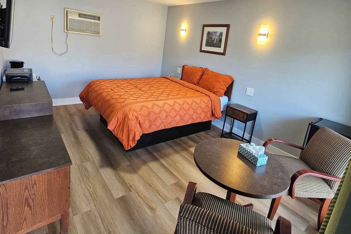 A room with a bed, a desk, a round table, and two chairs. There is a wall-mounted air conditioner and a picture on the wall.
