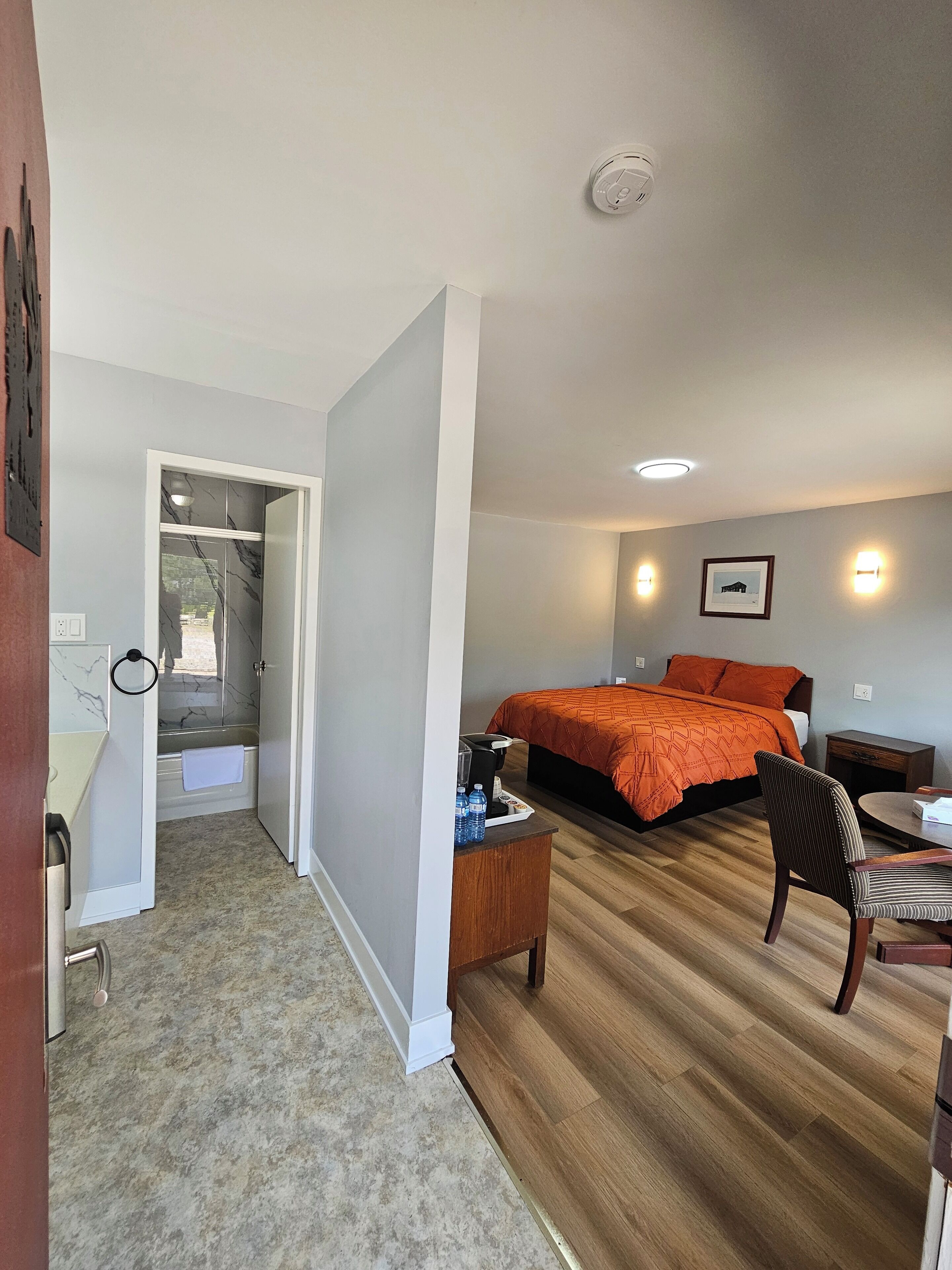 Habitación de hotel con cama, una silla, una mesita, mesita de noche, zona de ducha y baño.