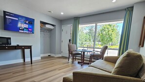 A spacious living area with a flat-screen TV mounted on the wall, a wooden floor, a sofa, a dining table with chairs, and a large window with curtains. - Arrowhead Inn by Baaman's (Huntsville)