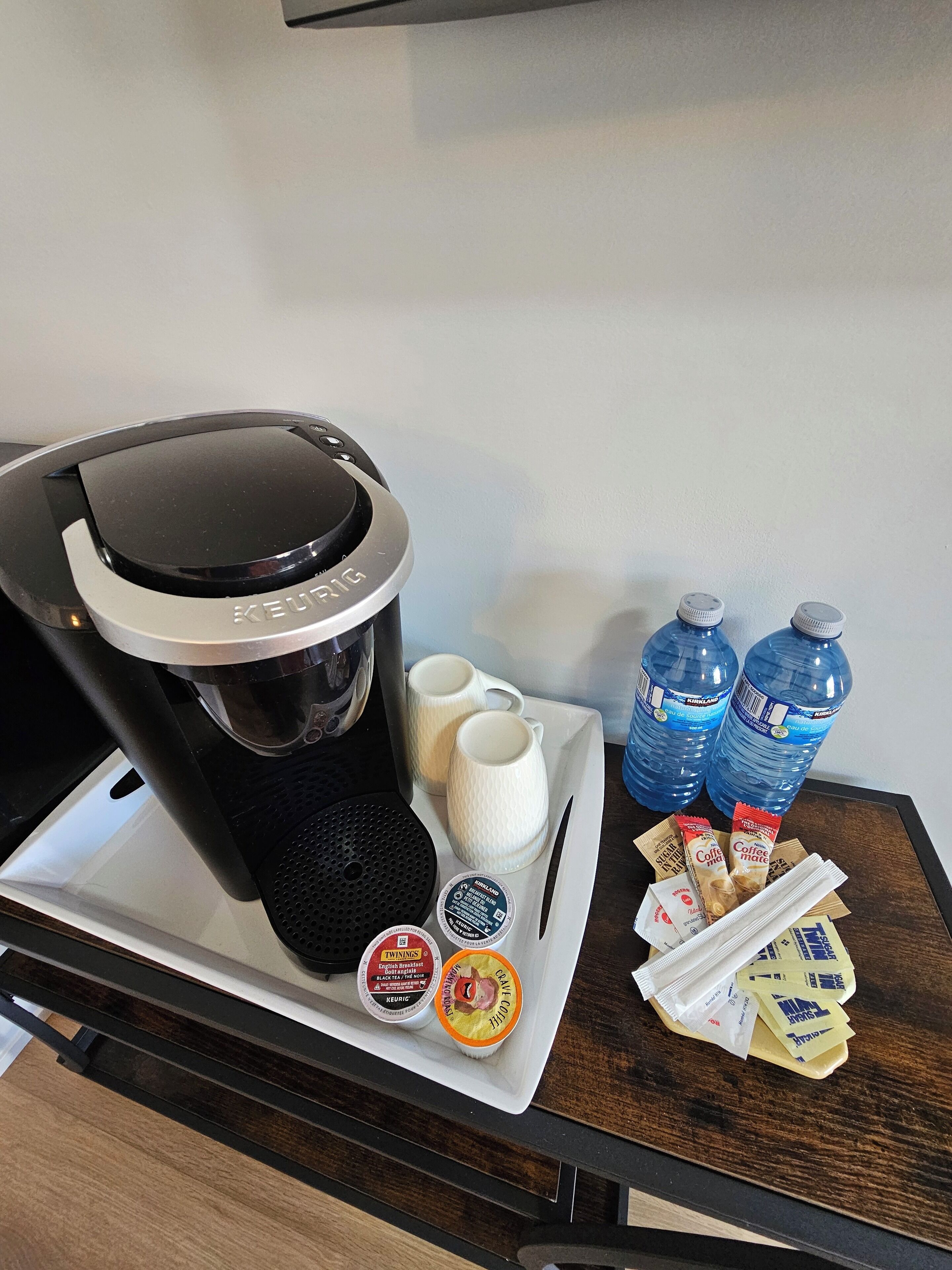 Une machine à café Keurig posée sur un plateau, accompagnée de deux tasses blanches, de deux bouteilles d’eau et de capsules de café.