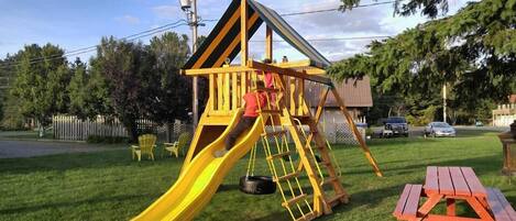 A playground with a yellow slide and climbing structure, surrounded by grass and trees.
