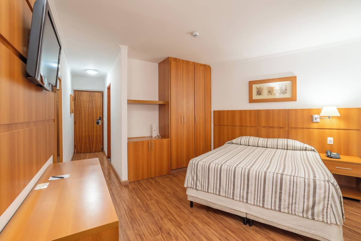 Una habitación de hotel con cama, armario de madera, escritorio, televisor montado en la pared y un cuadro enmarcado sobre la cama.