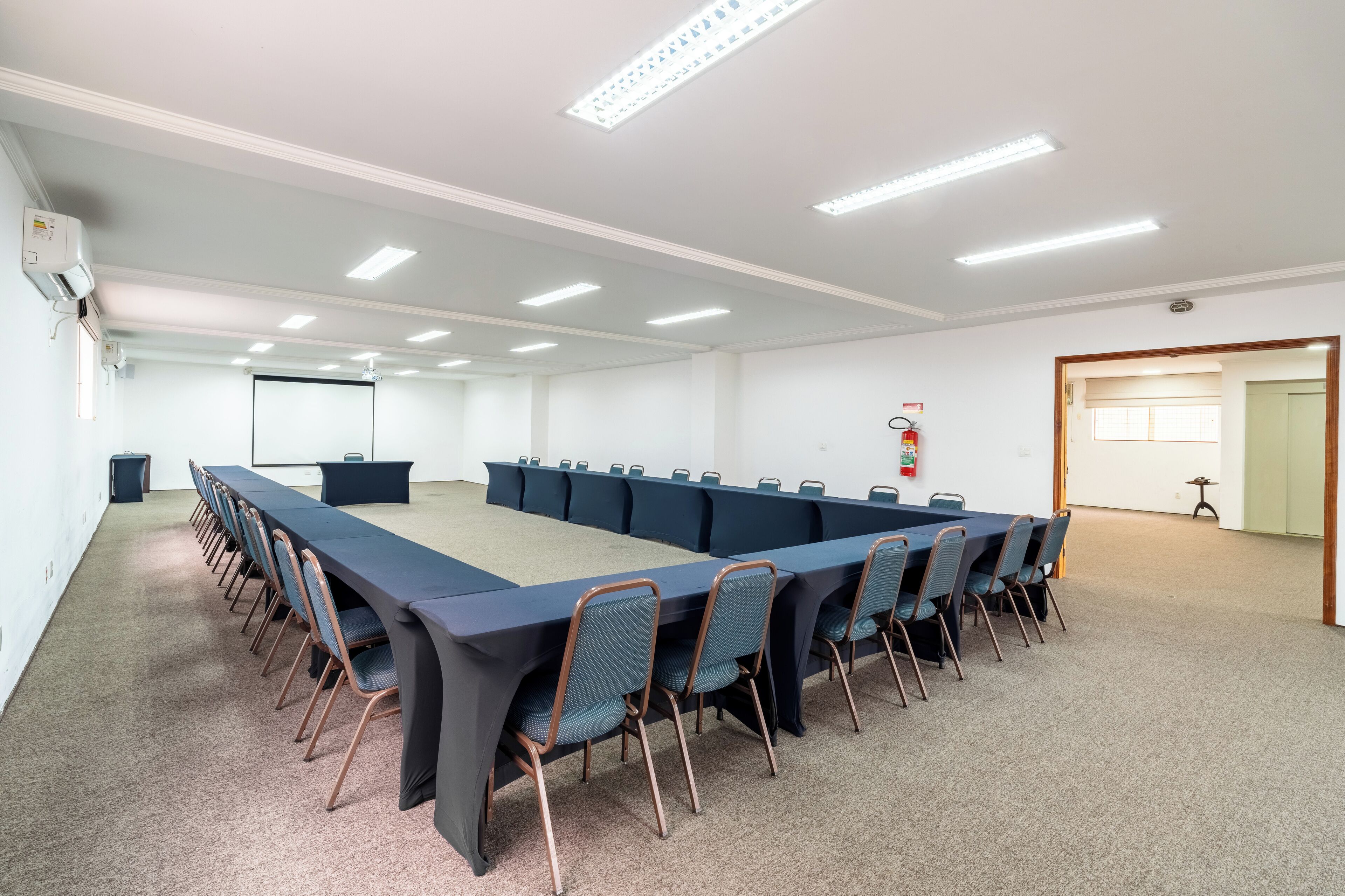 A conference room with a long table, chairs, a projector screen, and a fire extinguisher.