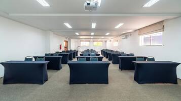 Un salón de conferencias con filas de mesas y sillas, un proyector y ventanas con persianas.