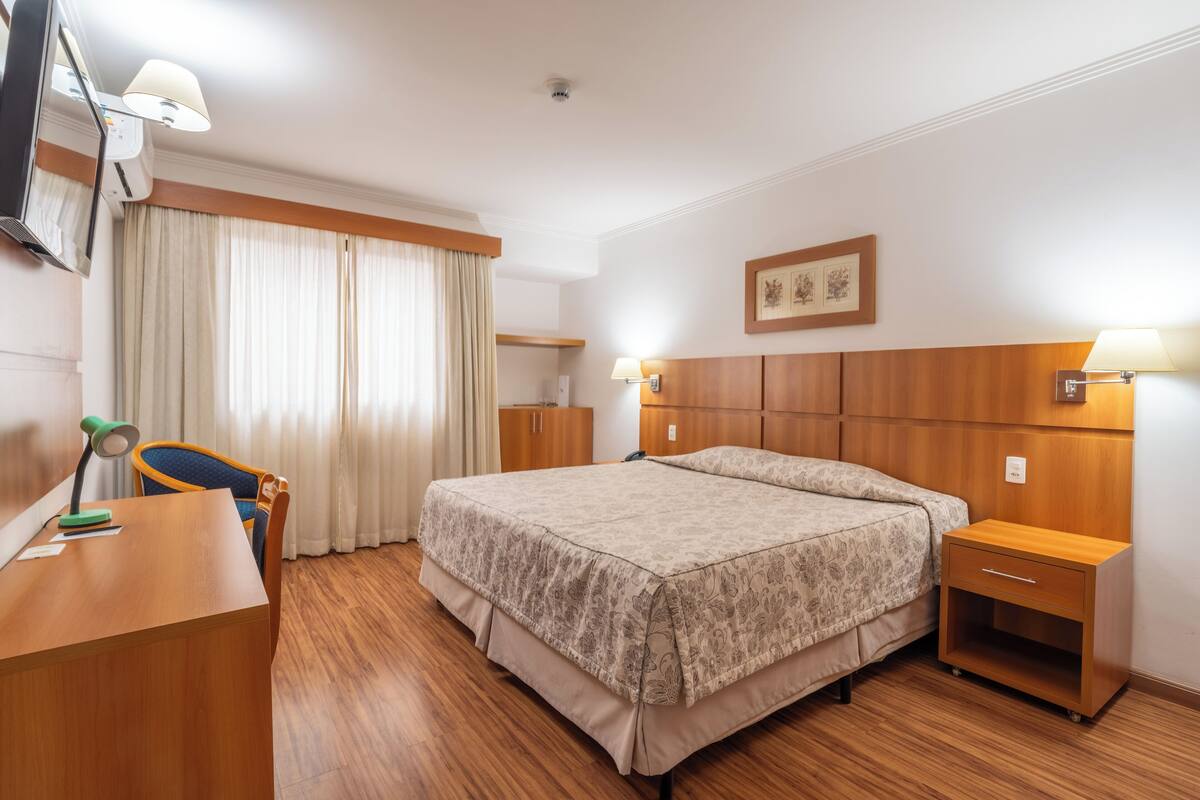 Habitación de hotel con cama de madera, escritorio con lámpara, silla, mesita de noche y televisor montado en la pared.