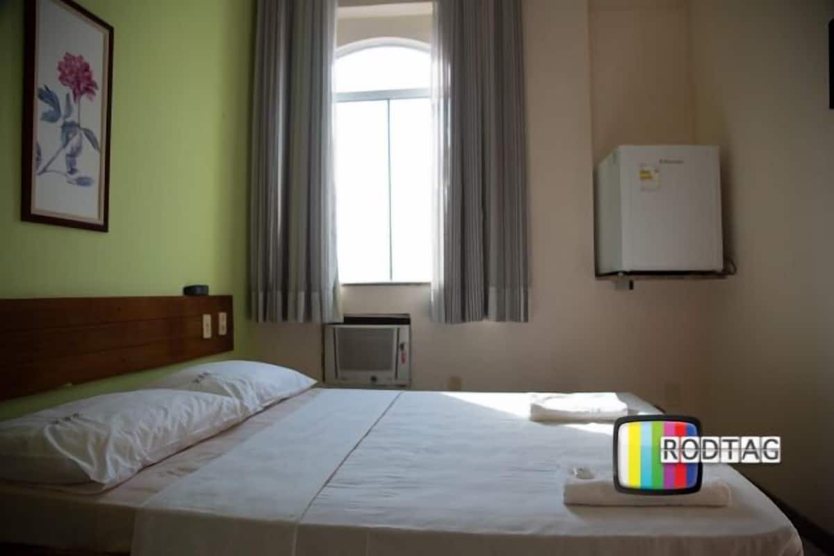 Une chambre d’hôtel avec un lit, une fenêtre avec des rideaux, un climatiseur fixé au mur et un tableau encadré accroché au mur.