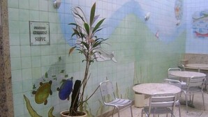 Paredes revestidas de azulejos com um mural, uma planta em vaso, cadeiras e mesas de metal em um ambiente.