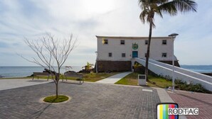 Edifício com fachada branca e porta azul, situado em área pavimentada com palmeira e bancos.