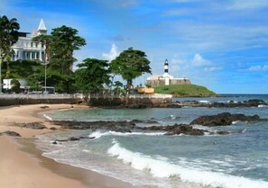A coastal scene with a lighthouse, a large building, and a beach. - Hotel Porto da Barra (Salvador)