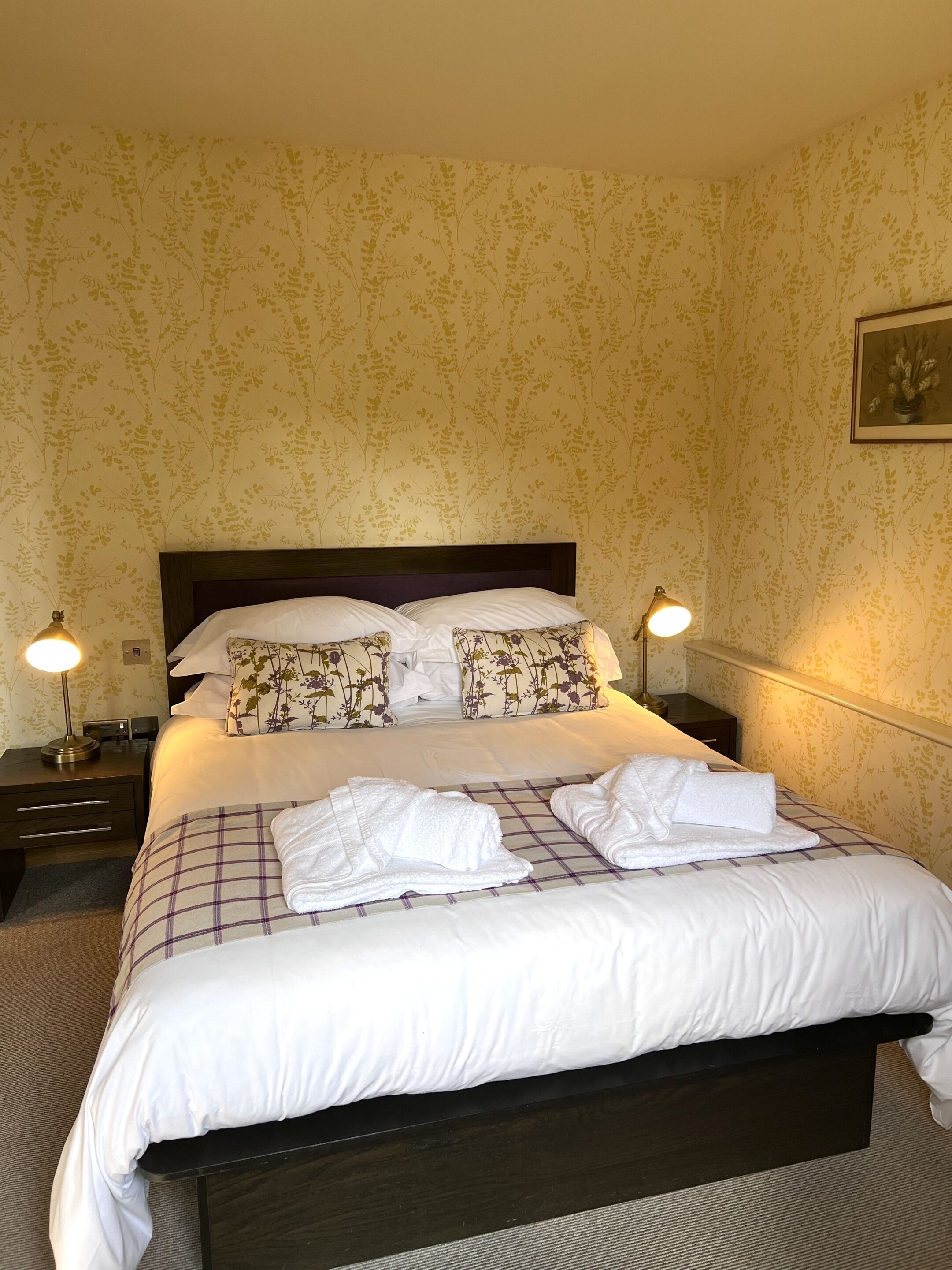 A neatly made bed with white linens and floral-patterned pillows, two bedside lamps, and a framed picture on the wall.
