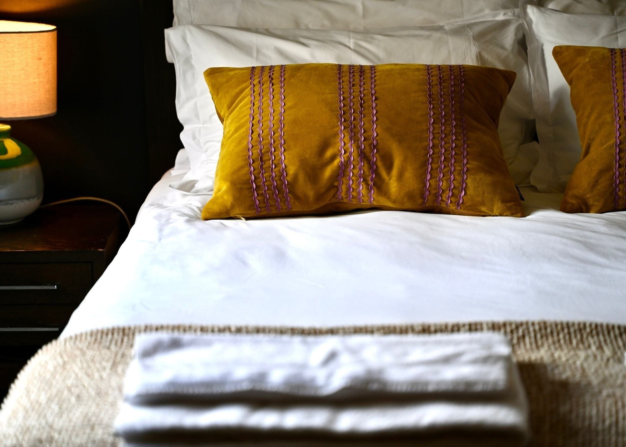 A neatly made bed with white linens and two mustard-colored pillows with purple stitching.