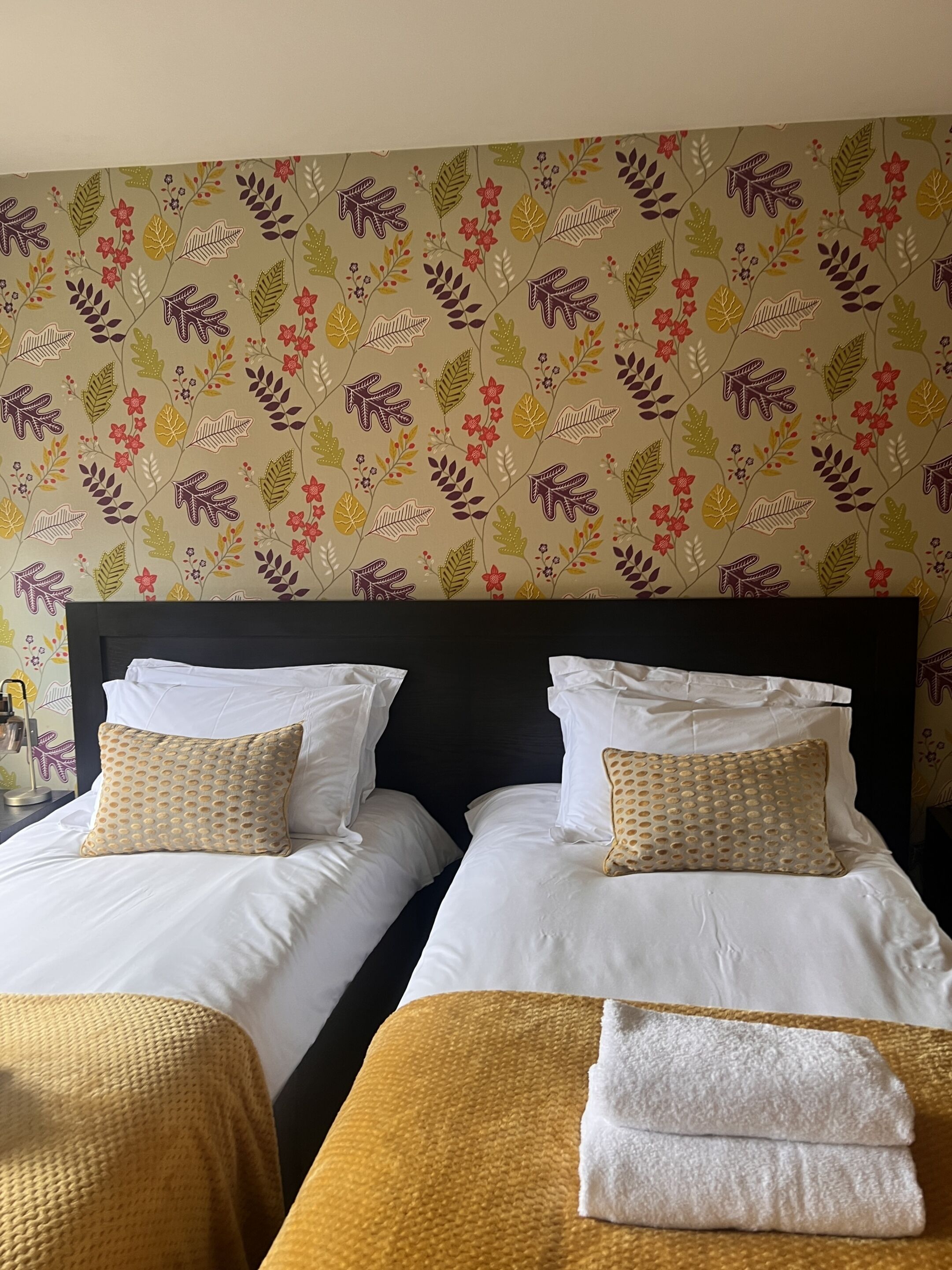 A hotel room with two beds, each with white linens and patterned pillows. The room has a dark headboard and a floral wallpaper design.