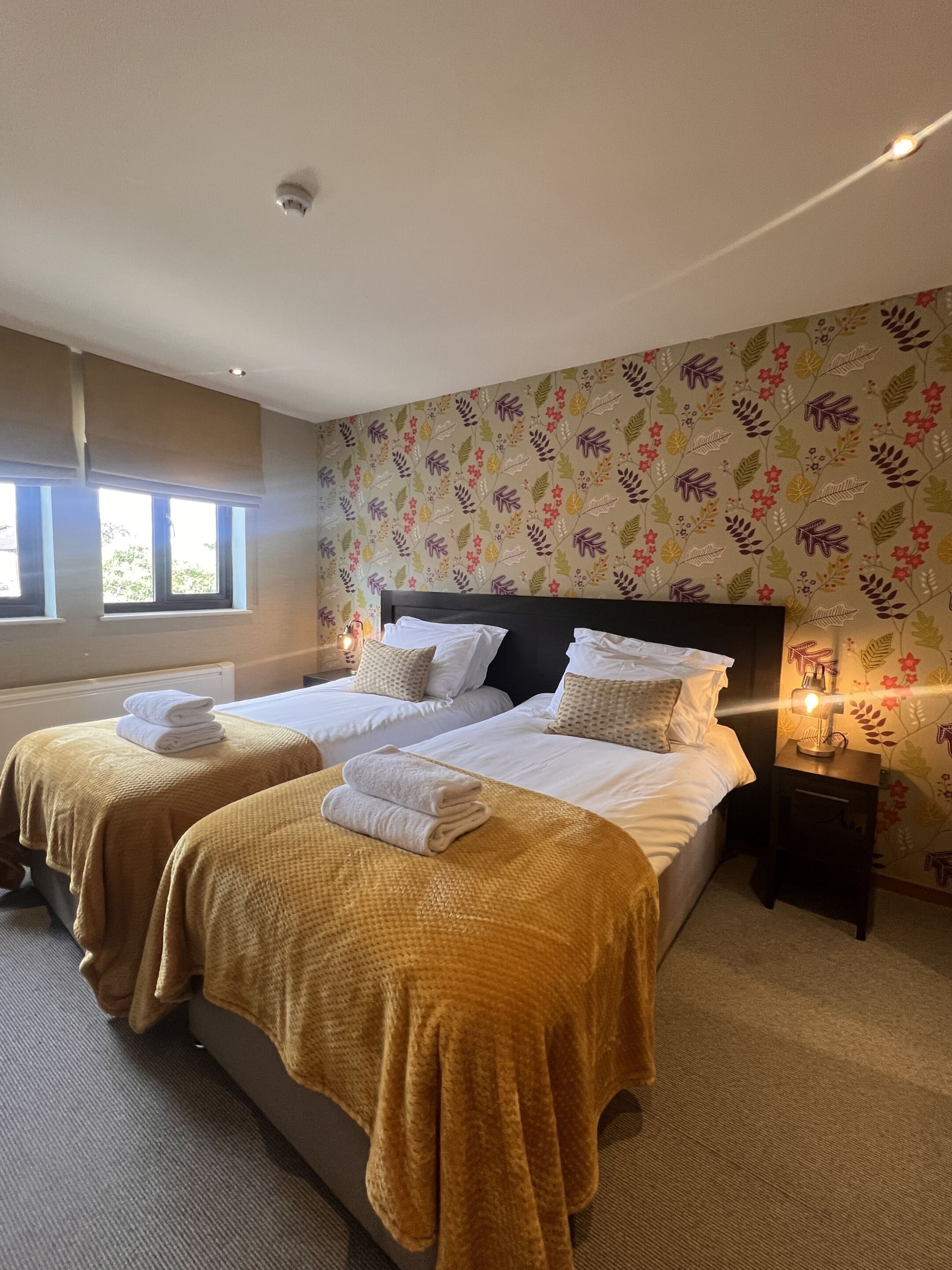 Habitación de hotel con dos camas, cada una con cubrecama amarillo y ropa de cama blanca. Se observa una ventana con persianas, una mesita de noche con lámpara y un papel tapiz estampado con motivos florales.