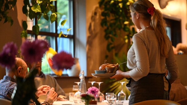 Ein Essbereich mit einem Tisch für zwei Personen, einem Kellner, der Speisen serviert, und einer Vase mit lila Blumen.
