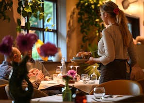 Ein Essbereich mit einem Tisch für zwei Personen, einem Kellner, der Speisen serviert, und einer Vase mit lila Blumen.