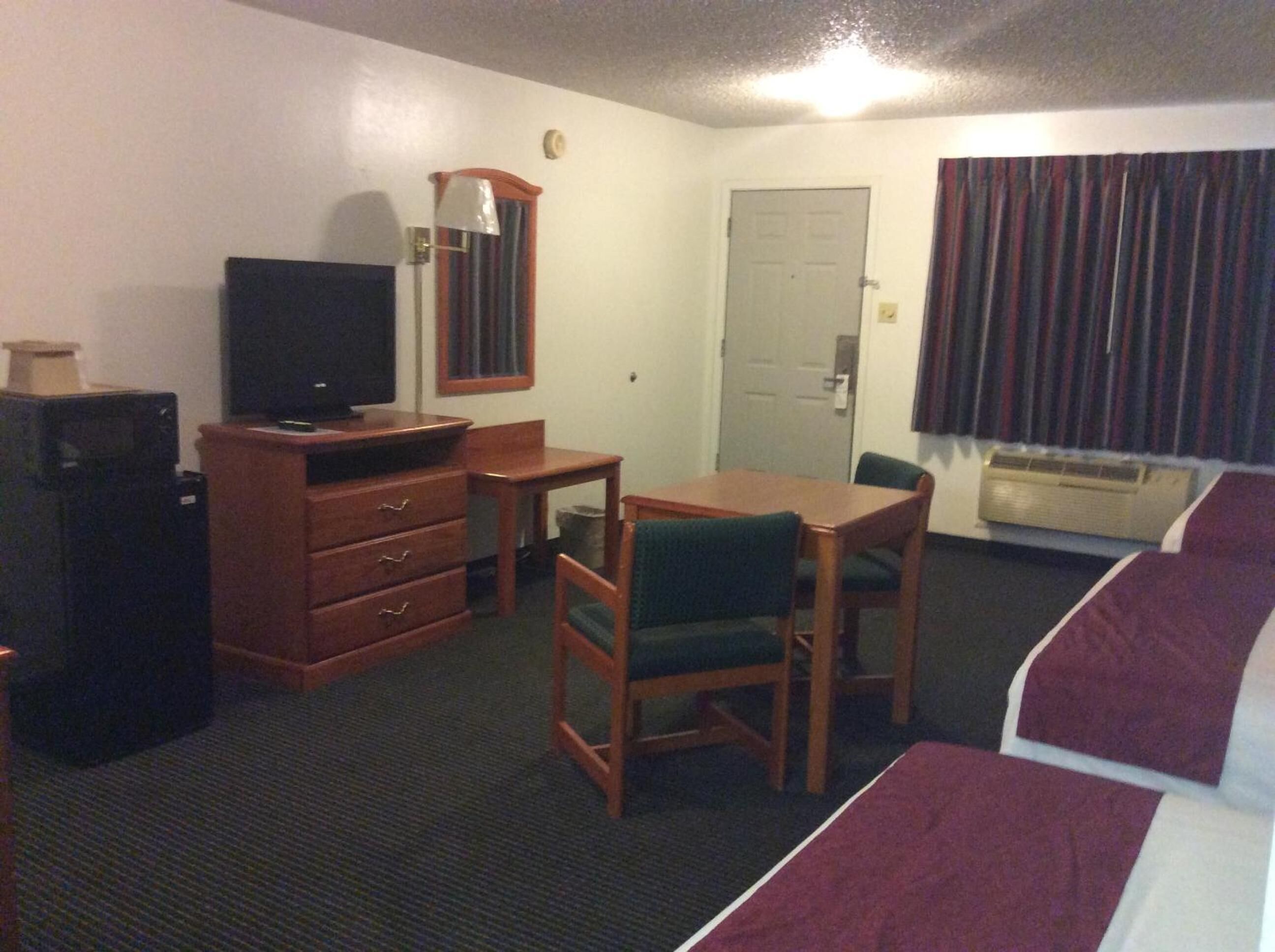 Una habitación de hotel con cama, escritorio, silla, televisor, cómoda y ventana con cortinas.