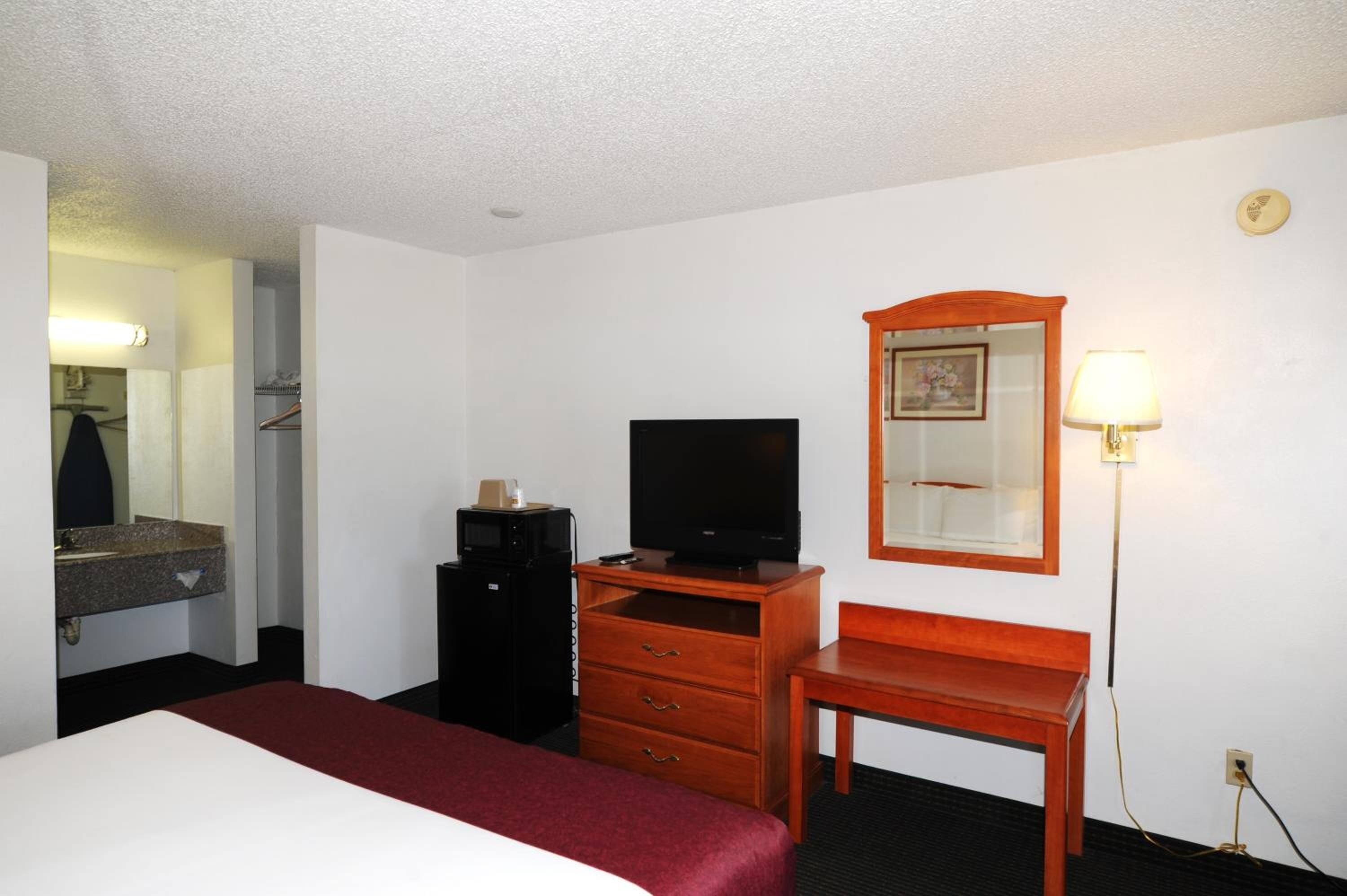 Una habitación de hotel con cama, televisor sobre un soporte de madera, escritorio, espejo y lámpara.