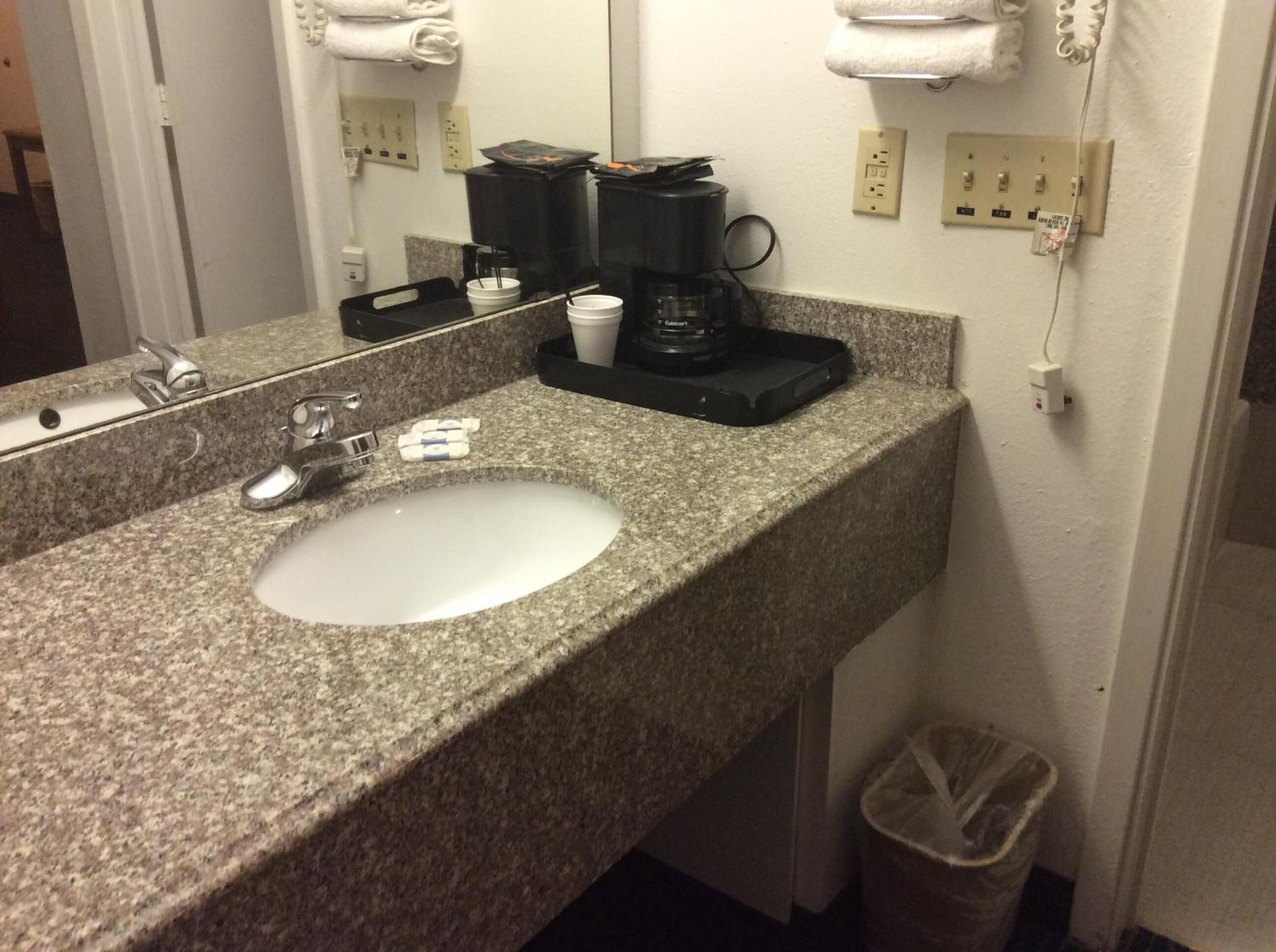 A hotel bathroom with a granite countertop sink, a mirror, a coffee maker, and a trash can.