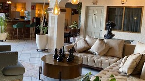 A spacious living area with a sofa, coffee table, and chairs. There are hanging lights, potted plants, and a view of the kitchen in the background. - Hotel GYA Express (Aguascalientes)