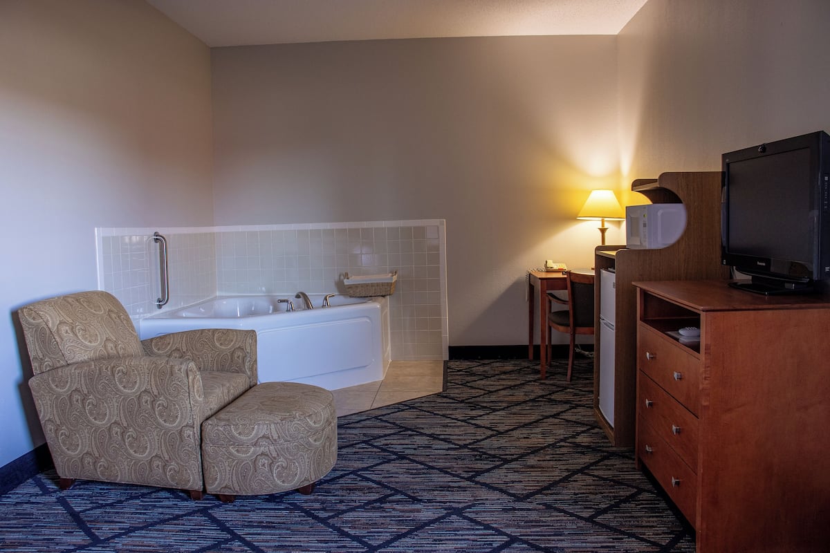 A hotel room with a bathtub, a chair, a wooden dresser, a television, and a lamp.