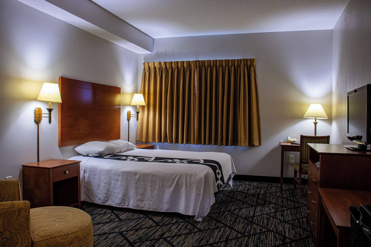 A hotel room with two beds, a wooden headboard, a large window with curtains, two bedside tables with lamps, a desk with a chair, and a patterned carpet.