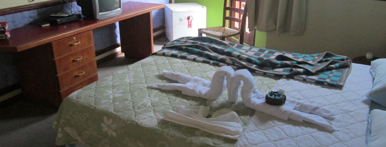 A hotel room with a bed, a wooden desk with drawers, a television, and a refrigerator.