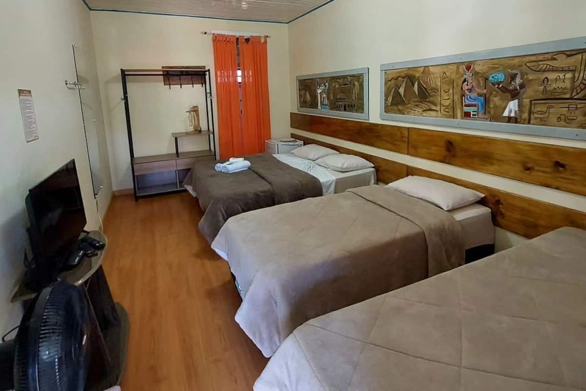 A hotel room with three beds, a desk with a computer and fan, a TV, and a window with orange curtains.