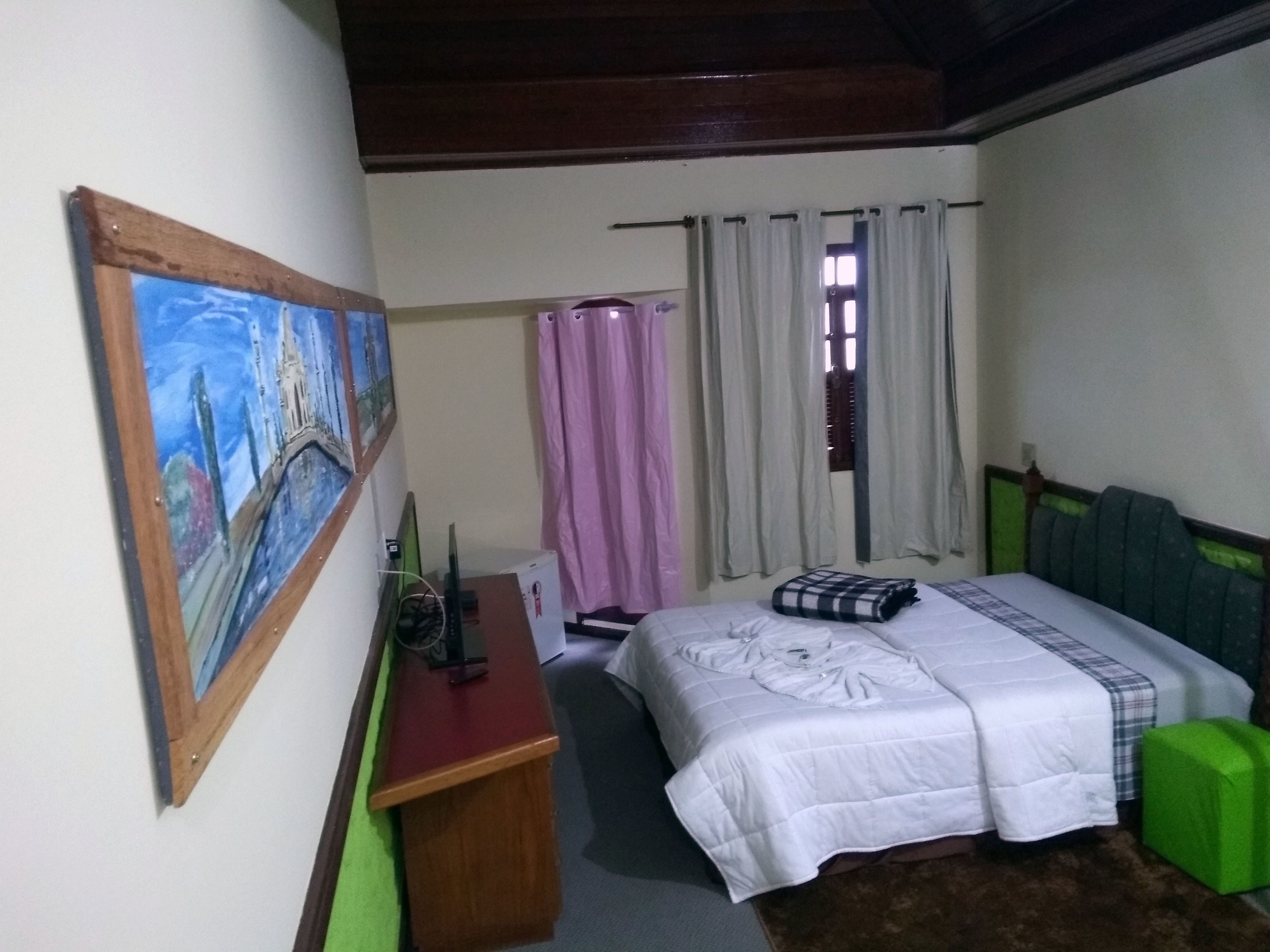 A bedroom with a bed, a desk, a chair, a painting, and a window with curtains.
