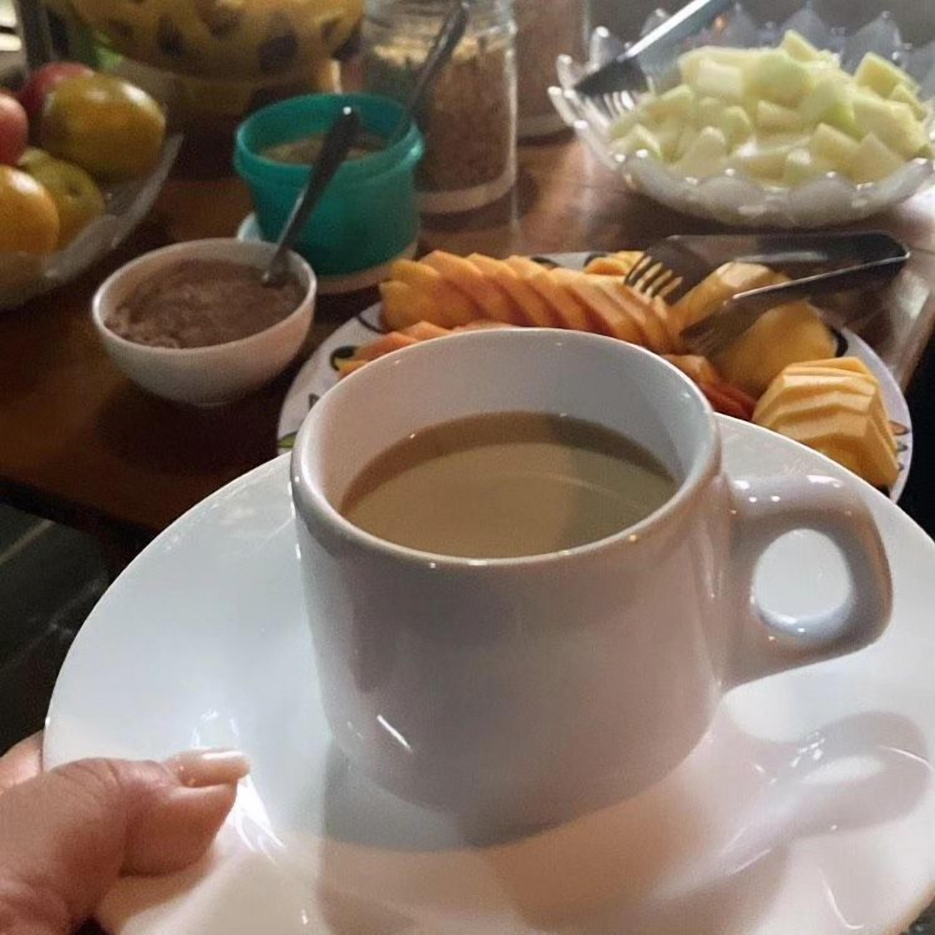 Una taza de café sobre un platillo, un bol de fruta en rodajas y un plato con pasteles sobre una mesa.
