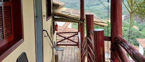 Um espaço coberto ao ar livre, com uma cadeira, uma mesa e vista para a paisagem, além de um corrimão de madeira.