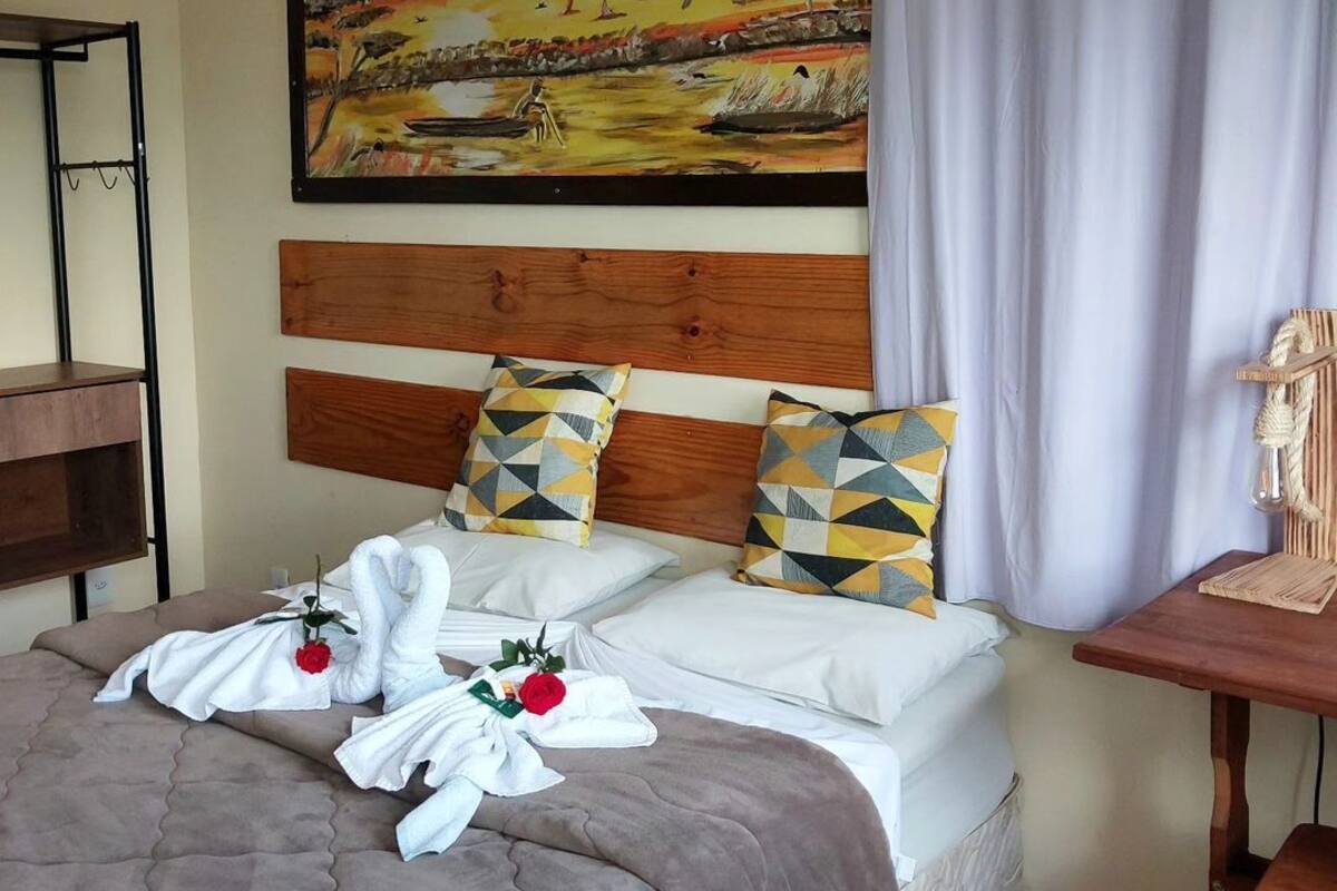 A hotel room with two beds, a wooden headboard, geometric patterned pillows, a painting on the wall, and a small table with a lamp.