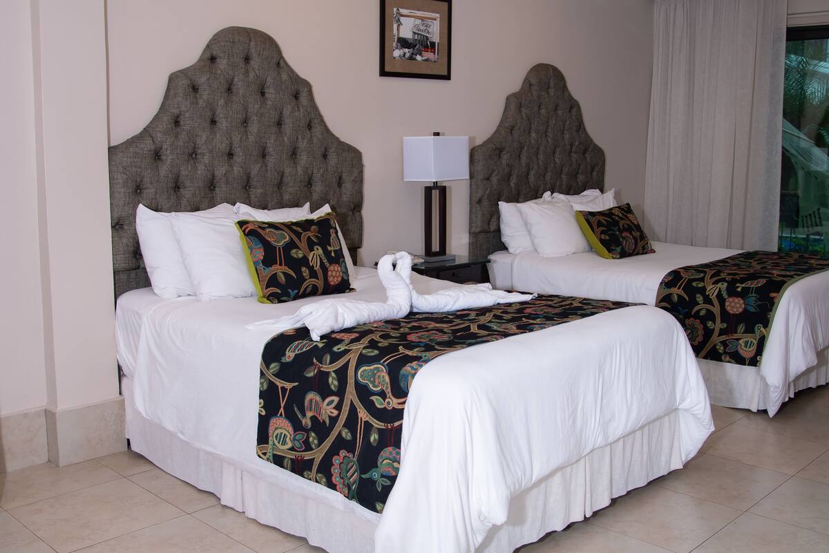 A hotel room with two beds, each with a tufted headboard, white bedding, and decorative pillows. A framed picture hangs on the wall above the beds.