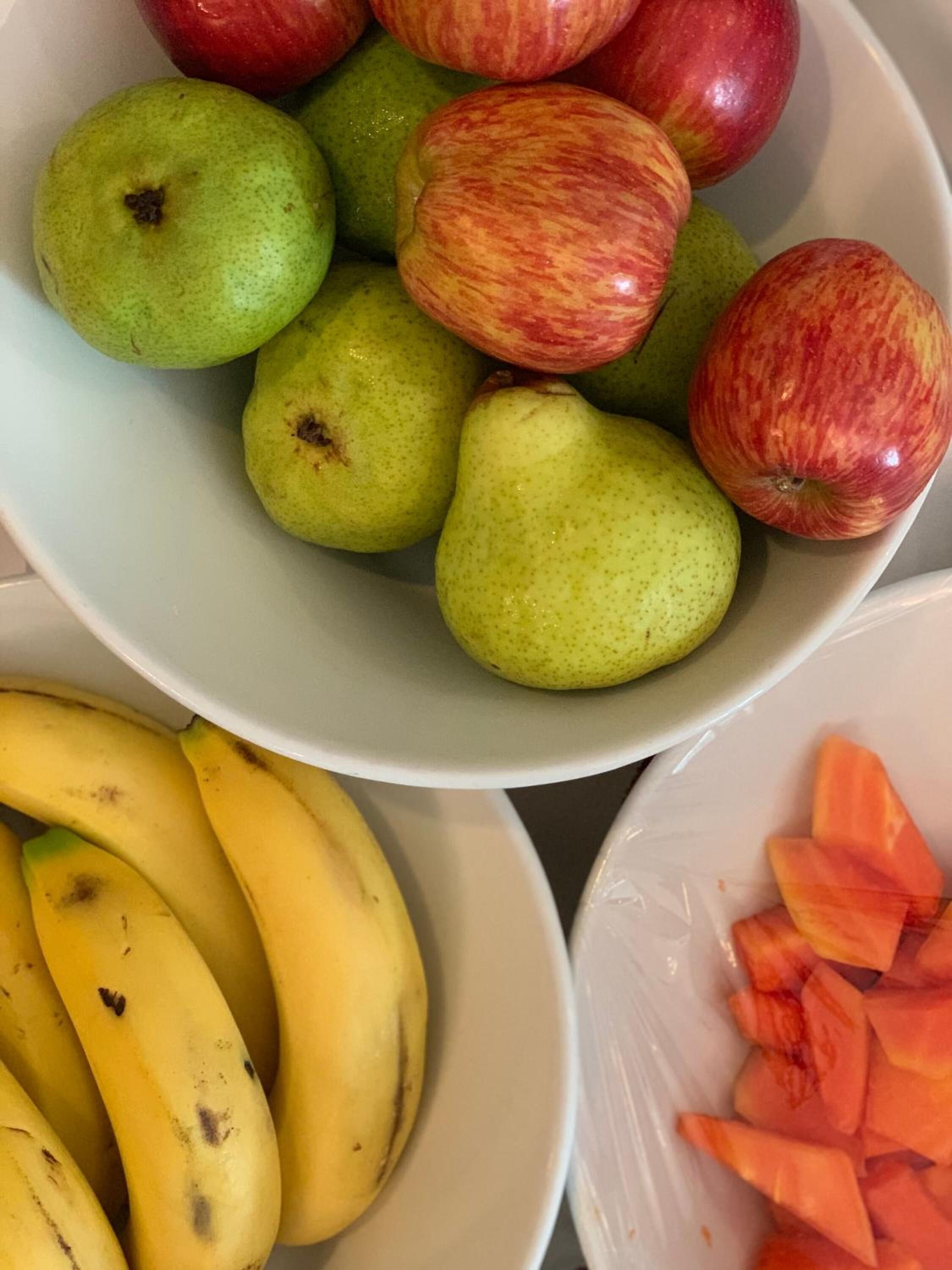 A bowl of green pears and red apples, a plate of bananas, and a bowl of sliced carrots.