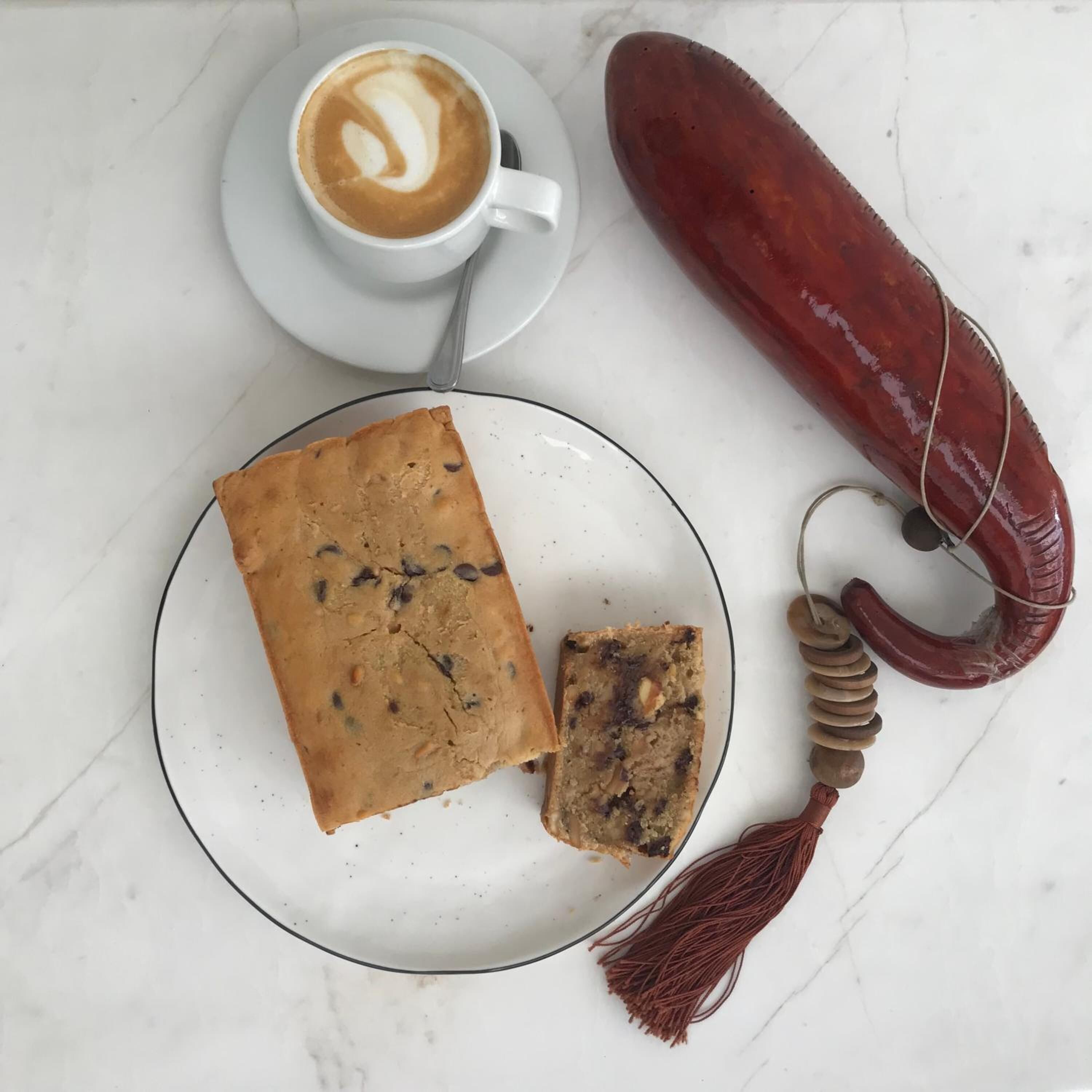 Un plato con un pan, una taza de café, una salchicha roja y un fleco decorativo.