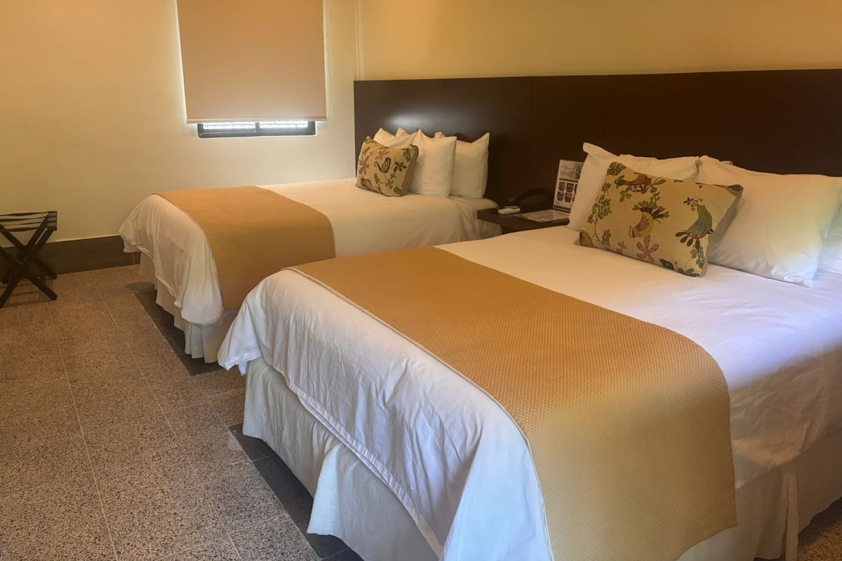 A hotel room with two beds, a headboard, a window with blinds, a small table with a phone, and a wall-mounted air conditioner.