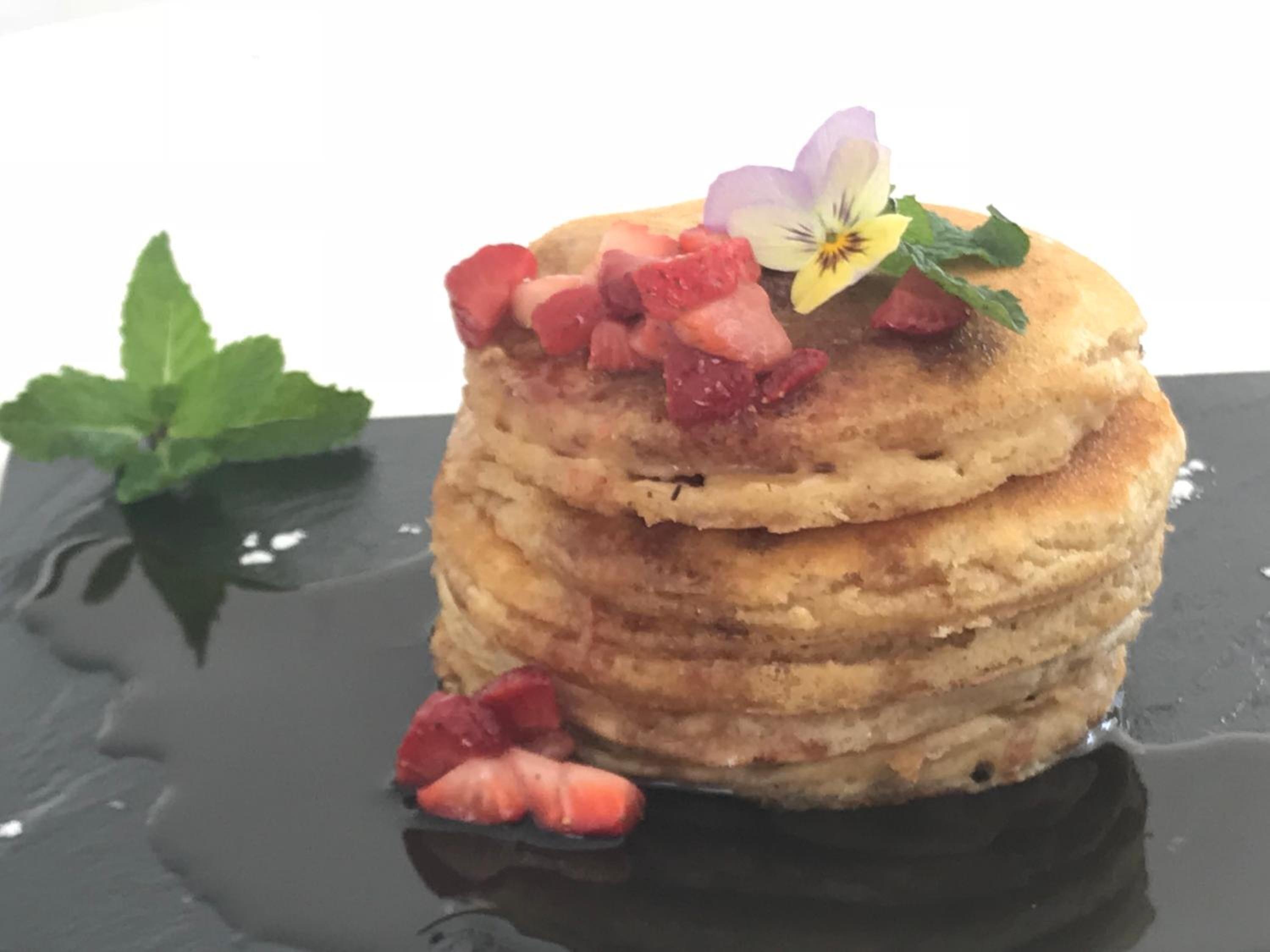 A stack of pancakes with strawberries and a flower on top, placed on a reflective surface.