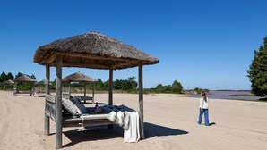 Una playa con un quiosco de techo de paja, una silla de descanso con una manta, y una persona de pie cerca.