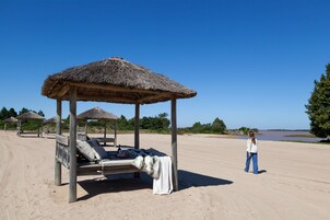 Una playa con un quiosco de techo de paja, una silla de descanso con una manta, y una persona de pie cerca.