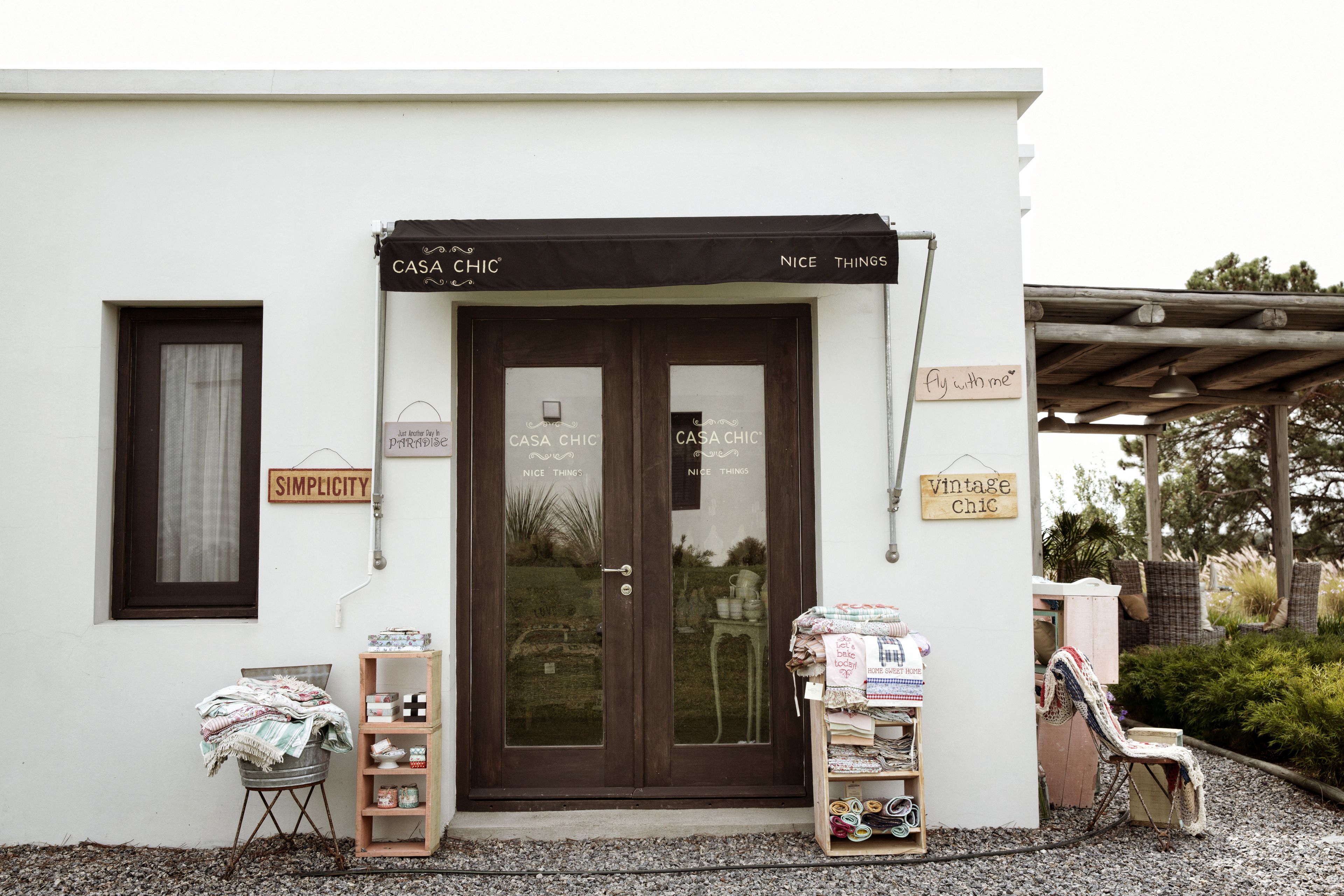 A storefront with a sign reading "CASA CHIC" and "NICE THINGS." There are two wooden doors, a small table with items, and a chair outside.