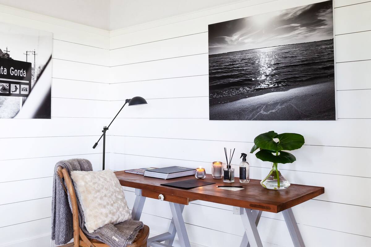 A room with a wooden desk, a chair with a cushion, a plant, and a framed picture of a beach.