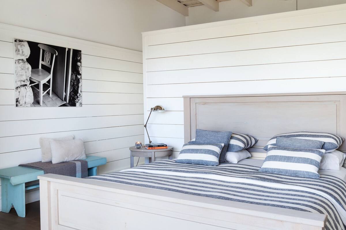 A bedroom with a wooden ceiling, a bed with striped bedding, a nightstand with a lamp, and a patterned rug.