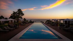 Vista de la piscina con sillones y sombrillas, con vista a un cuerpo de agua al atardecer.