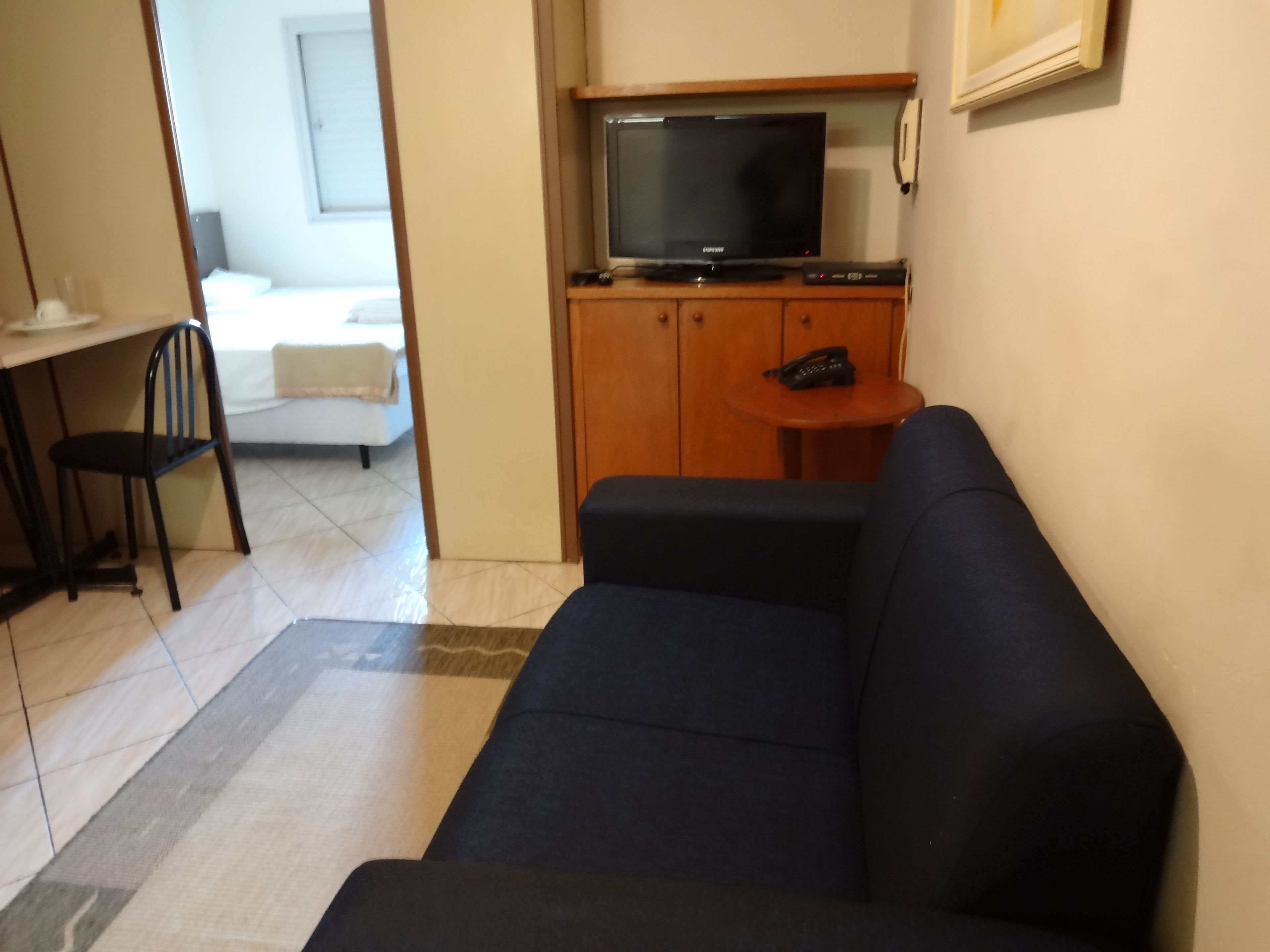 Un salon avec un canapé foncé, un téléviseur posé sur un meuble en bois, une petite table ronde avec un téléphone et un fauteuil.