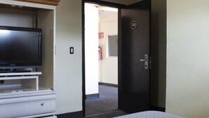 Una habitación de hotel con una cama, un televisor sobre un mueble, una puerta con un número y un pasillo que da a otra habitación.