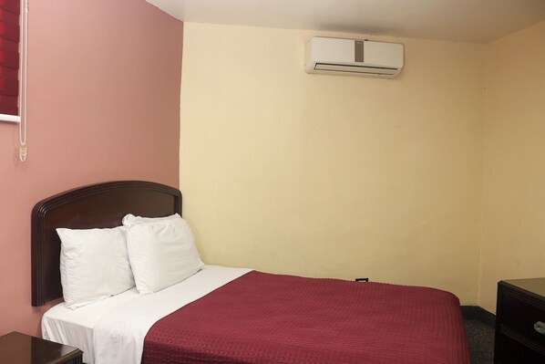 Una cama individual con ropa de cama blanca y roja, un cabecero de madera, una mesita de noche y una pequeña veladora.