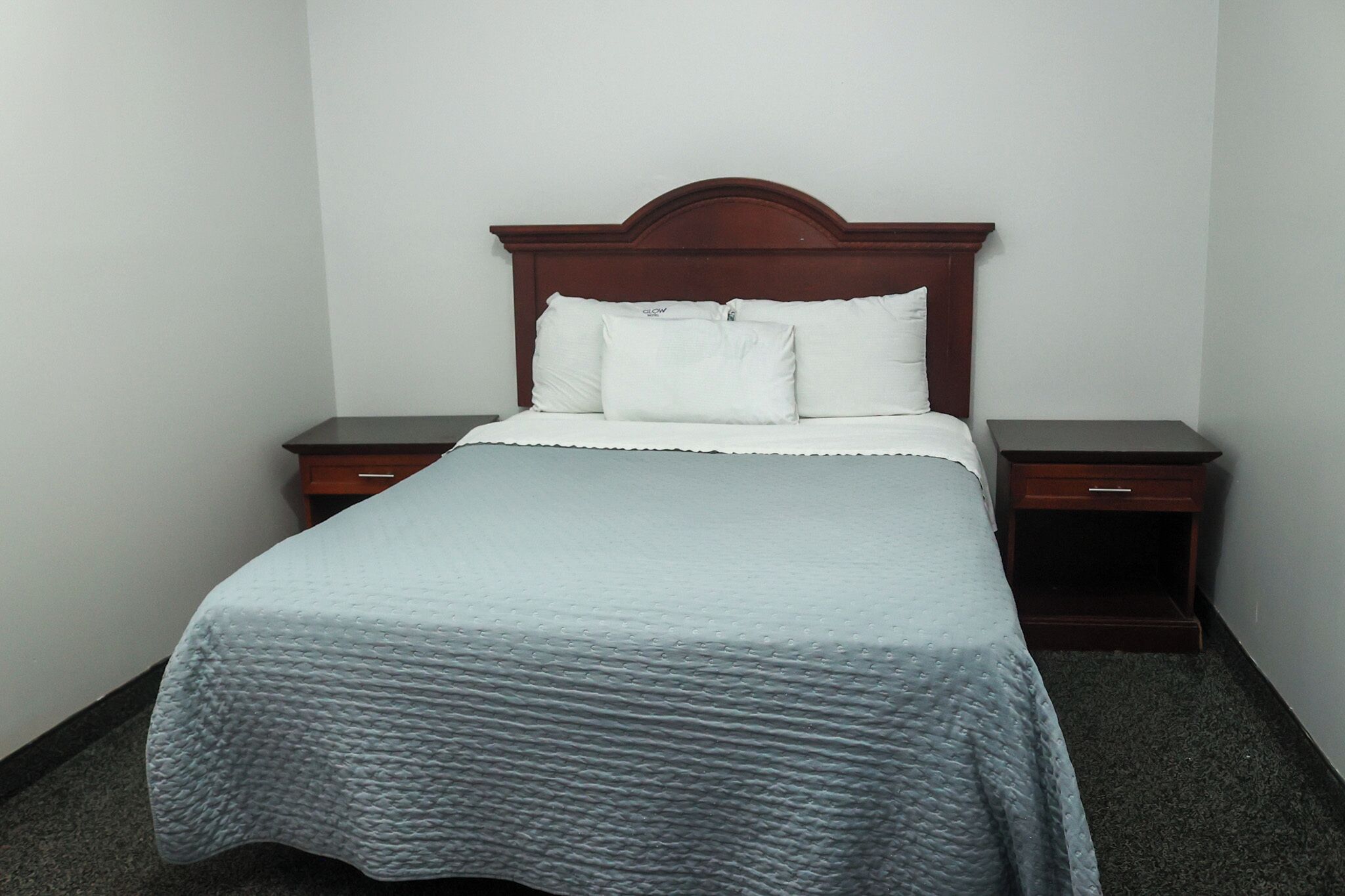 A neatly made bed with a wooden headboard, two bedside tables, and a plain wall.