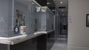 Zona de recepción de hotel con un separador de vidrio, una pantalla digital y un cartel de prohibido fumar.